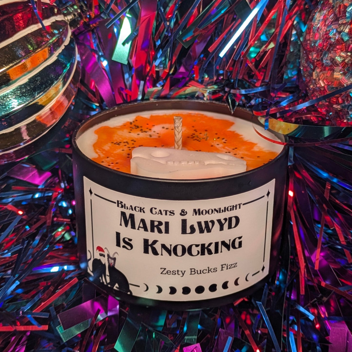 Candle with a label 'Mari Lwyd Is Knocking' surrounded by festive tinsel.