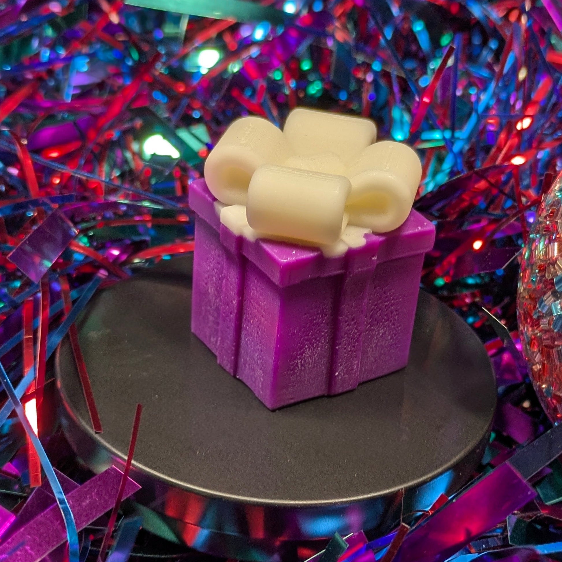 A wax melt shaped like a purple gift box with a white bow with colorful, festive background