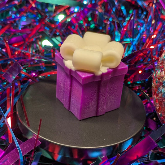 A wax melt shaped like a purple gift box with a white bow with colorful, festive background